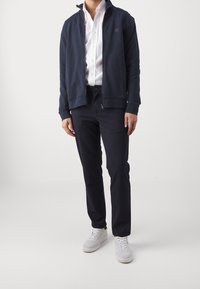 Marinblå zip-jacka i mjukt tyg, ihopparad med åtsittande mörka byxor och vita sneakers. Skjortan är vit med knappar och visar en minimalistisk stil.