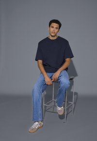 Mladý muž s kudrnatými vlasy, oblečený v tmavém volném tričku, modrých džínech a béžových teniskách, sedící na vysokém kovovém stoličce na šedém pozadí.