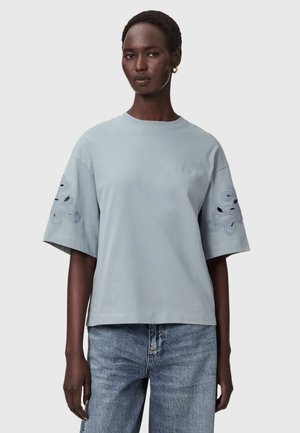 Kvinde iført lyseblå oversized T-shirt med blomsterbroderi på ærmerne og lyseblå denimjeans, stående foran en ensfarvet baggrund.