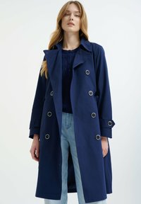 Granatowy płaszcz trench w lekkim materiale, podwójnie zapięty design z metalowymi guzikami, duży kołnierz i regulowane mankiety. Dopasowany krój.