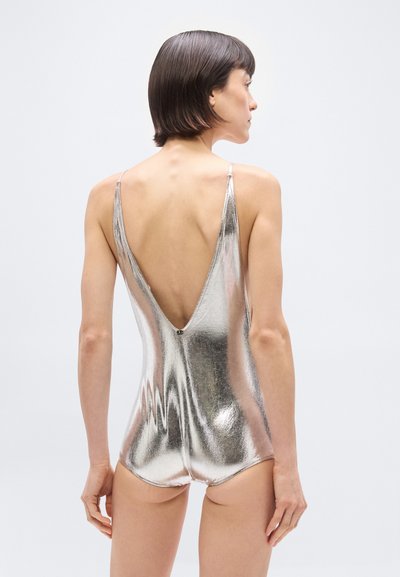 Γυναίκα με κοντά σκουρόχρωμα μαλλιά, φορούν μια λαμπερή ασημένια μεταλλική φόρμα μπικίνι με βαθιά λαιμόκοψη στο πίσω μέρος, στέκεται μπροστά σε ένα απλό φωτεινό φόντο.