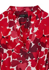 Blusa de estampado floral rojo con escote en V. Presenta grandes flores rojas y burdeos oscuros sobre un fondo blanco. Tejido ligero. Etiqueta de marca visible.