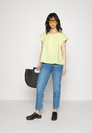 Top de mangas curtas verde-lima com detalhes em botão, jeans de denim azul, clogs pretos com detalhes em tachas e uma bolsa tecida preta texturizada.