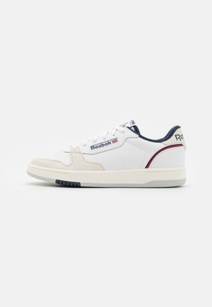 Sneaker basse Reebok bianche con dettagli navy e rossi, punta in suede e logo sul lato e sul tallone su sfondo semplice.