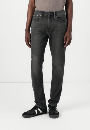 Jeans de mezclilla negra con un corte recto, cinco bolsillos y costuras en contraste. Llevados con zapatillas negras que tienen acentos en blanco.