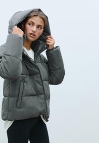 Puffer jacket in dark gray, featuring a zip closure, large hood, and quilted texture. Two side pockets and relaxed fit design.