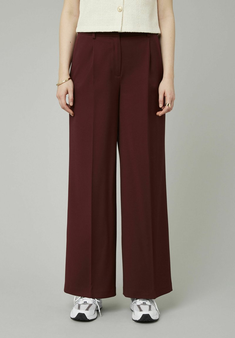 Pantalons larges en tissu bordeaux profond, dotés de plis avant, d'une coupe ajustée et d'une texture lisse, associés à des baskets blanches.