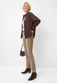 Brown button-up shirt over a beige turtleneck, paired with light brown pants and dark brown ankle boots. Small handbag at the side.
