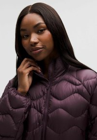 Fioletowa kurtka puchowa z wysokim kołnierzem, z pikowanym falistym wzorem i zamkiem błyskawicznym z przodu. Gładka, błyszcząca tekstura.