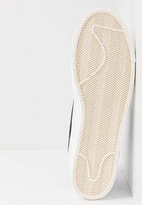 Sneaker bianca con suola in gomma testurizzata caratterizzata da un motivo a spina di pesce, accenti neri lungo il lato e materiale liscio sulla parte superiore.