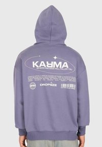 Person, die einen lila Kapuzenpullover trägt mit dem Text "KARMA Tu Gutes und Gutes wird dir folgen" und zusätzlichem kleinerem Druck auf dem Rücken.