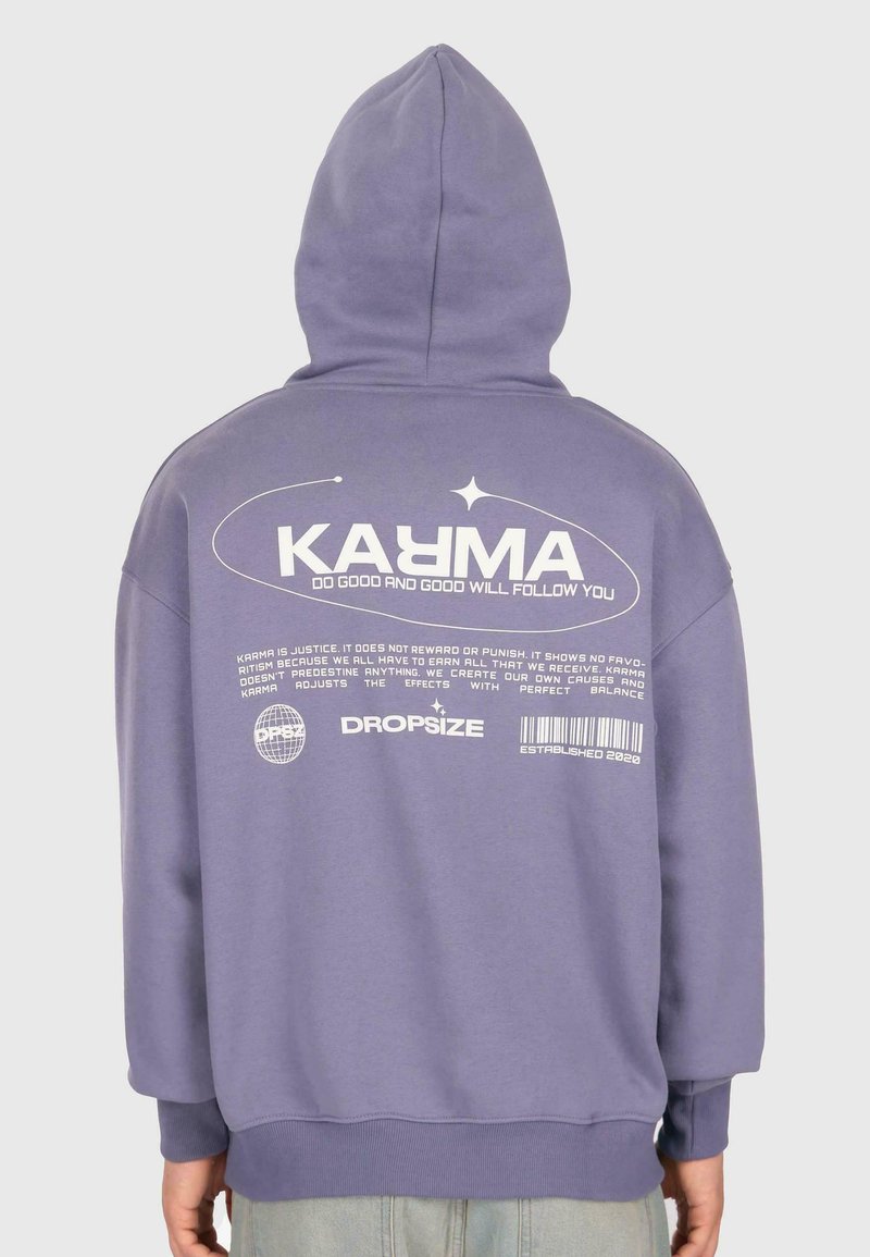Person, die einen lila Kapuzenpullover trägt mit dem Text "KARMA Tu Gutes und Gutes wird dir folgen" und zusätzlichem kleinerem Druck auf dem Rücken.