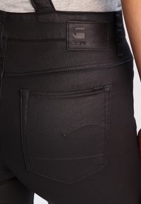 Zbliżenie na tylną kieszeń czarnych dopasowanych jeansów z wytłoczonym skórzanym naszywką G-Star RAW i przeszywanym logo.