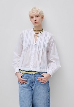 Person mit kurzen blonden Haaren trägt eine weiße Bluse mit Knöpfen, blaue Jeans, einen hellen Gürtel, einen Ring und gesteppte Halsketten und steht vor einer schlichten Wand.