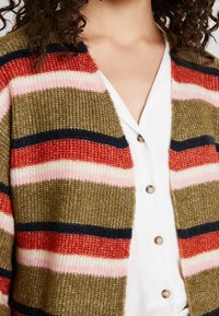 Pletený kardigan se zelenými, růžovými, červenými a námořnickými pruhy. Otevřený přední střih s měkkou texturou, nošený přes bílou košili na knoflíky.