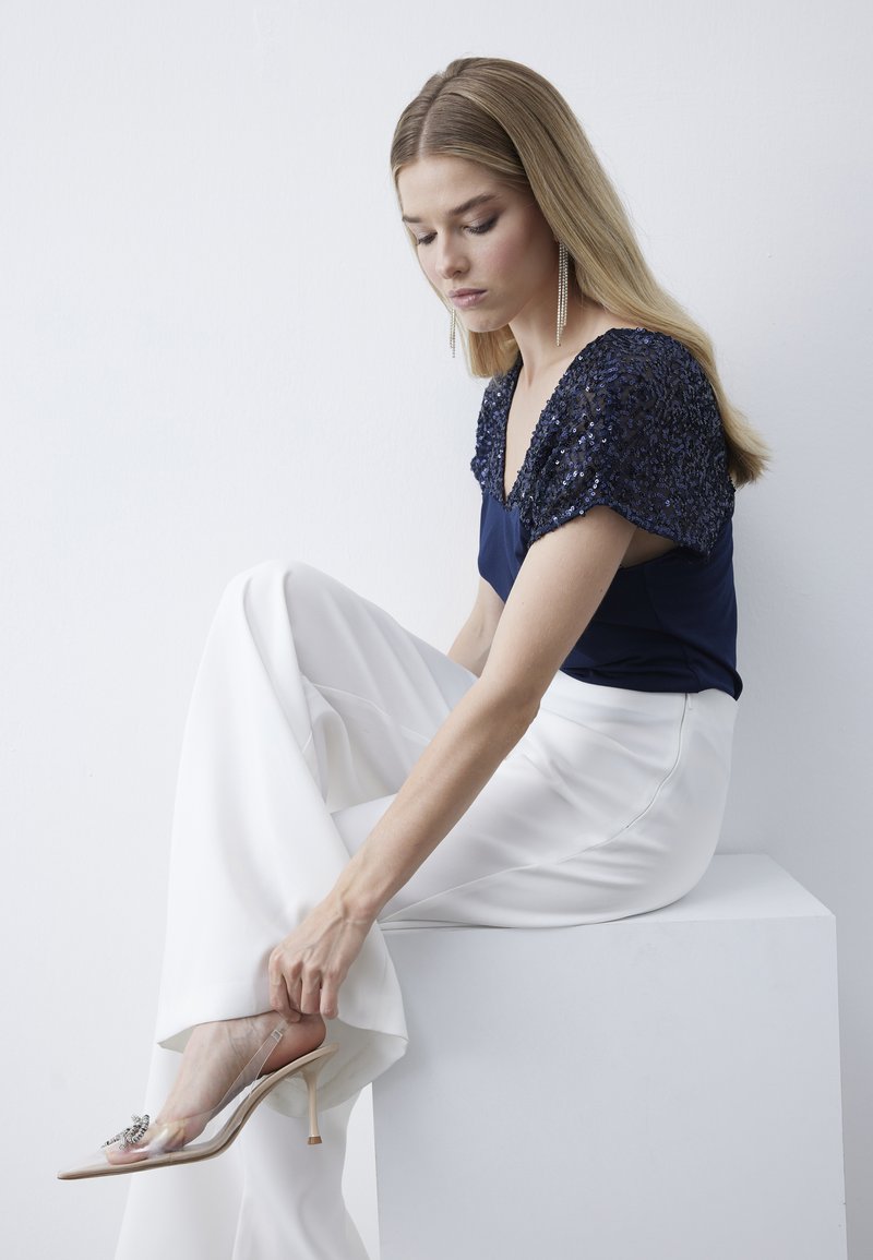 Top blu navy con maniche ricamate di paillettes, pantaloni bianchi a gamba larga e sandali trasparenti con tacco e decorazioni. Texture morbide e design moderno.