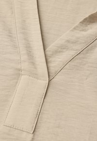 Beige tyg med en mjuk textur och subtilt rynkat mönster. Har en V-formad halslinning och synlig sömnad, vilket skapar en ren design.