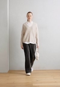 Suéter beige esponjoso con cuello en V sobre una camisa blanca con cuello, combinado con pantalones oscuros y zapatillas blancas. Lleva un bolso blanco.