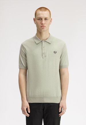 Ung mand med kort rødt hår iført lysegrå kortærmet poloshirt med krave og sorte bukser, stående mod en ensfarvet hvid baggrund.