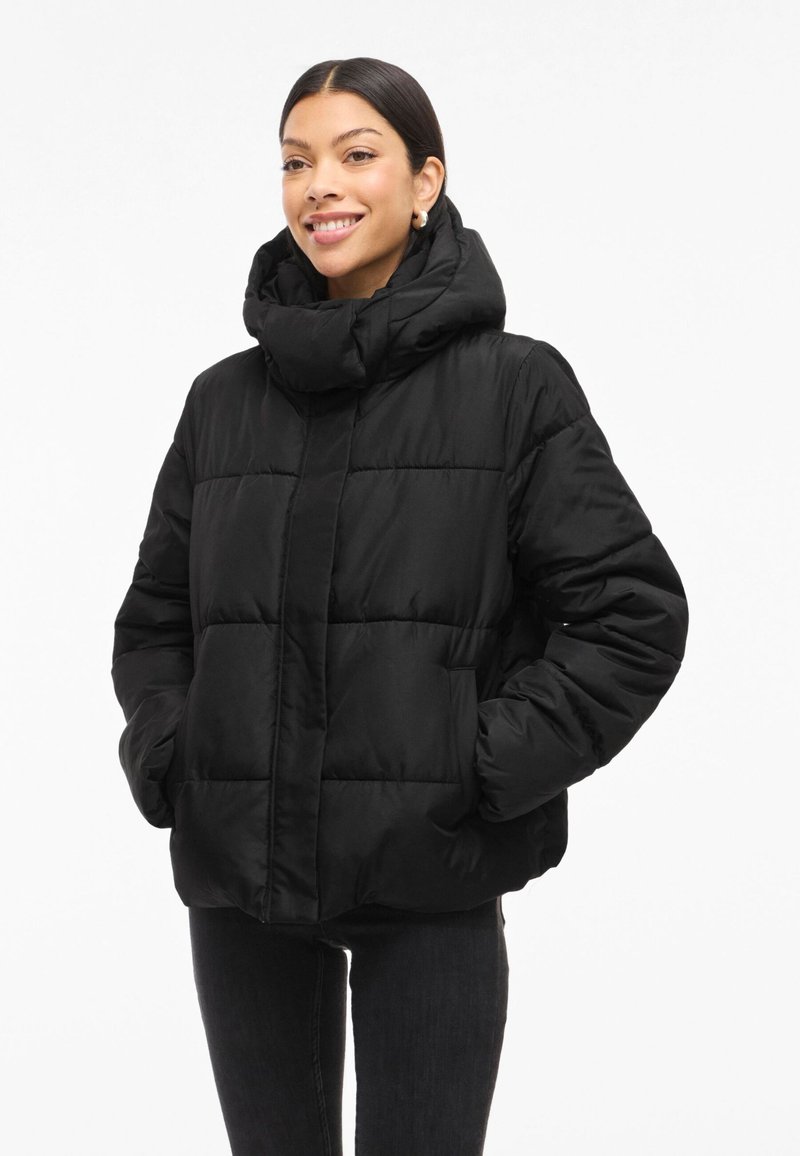 Casaco puffer preto com gola alta e capuz, apresentando um design acolchoado e dois bolsos frontais. Textura lisa e brilhante.