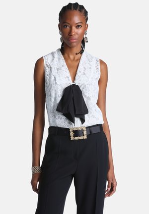 Frau trägt eine ärmellose weiße Spitzenbluse mit schwarzer Schleife, schwarze Hose, Gürtel mit goldener Schnalle, silberne Ohrringe und ein silbernes Armband.