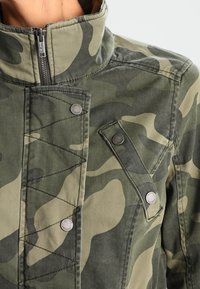 Bunda se vzorem maskáče v zelených tónech, s vysokým límcem, zipovým zapínáním a kovovými knoflíky s prošívaným designem.