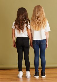 To børn holder hinanden i hånden, iført hvide T-shirts, sorte og blå stramme jeans. Sorte og hvide sneakers. Langt bølget hår, set bagfra.