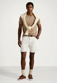 Καφέ κοντομάνικο πουκάμισο, λευκές τζιν βερμούδες με τριμμένες άκρες, κρέμα πουλόβερ δεμένο στους ώμους, καφέ αθλητικά παπούτσια και μπεζ κάλτσες.