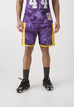 Mann trägt lila und gelbe Basketballshorts und passendes Trikot mit Drachenmustern, schwarze Socken und schwarze Sportschuhe.
