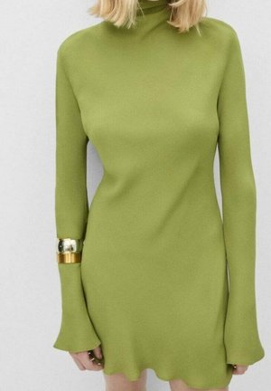 Kvinde iført en tætsiddende limegrøn kjole med lange ærmer, høj halsudskæring og flagrende manchetter, accessoriseret med to metallic armbånd.