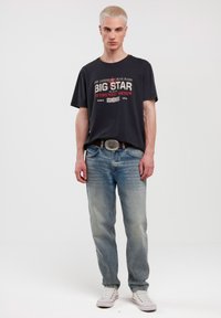 Crna majica s grafikom "BIG STAR" u bijeloj i crvenoj boji, plave izblijeđene traperice, srebrna kopča za pojas i bijele tenisice.