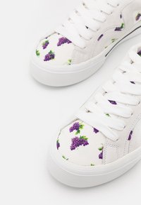 Weiße Leinenschuhe mit einem Muster von Weinreben in Lila und Grün. Gummisohle und weiße Schnürsenkel runden das Design ab.