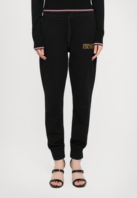Sorte joggebukser med snor i livet, ribbet mansjetter, og "VERSACE JEANS COUTURE"-logo i gull. Kombinert med sorte stroppesandaler.