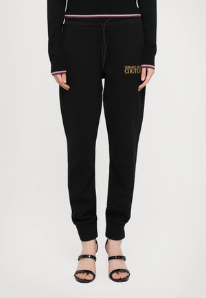 Pantaloni tuta neri con vita elastica e cordino, polsini a costine e logo "VERSACE JEANS COUTURE" in oro. Abbinati a sandali neri con lacci.