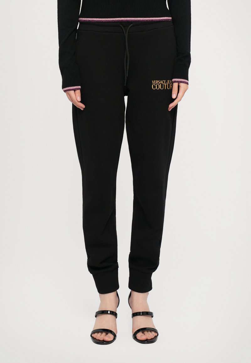 Sorte joggebukser med snor i livet, ribbet mansjetter, og "VERSACE JEANS COUTURE"-logo i gull. Kombinert med sorte stroppesandaler.
