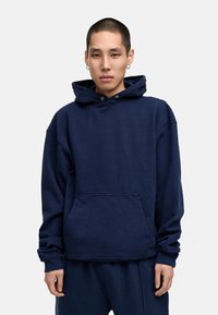 Marineblaue Kapuzenjacke aus Baumwolle, mit einer Fronttasche, Kordelzug-Kapuze und gerippten Bündchen. Lockerer Schnitt mit überschnittenen Schultern.