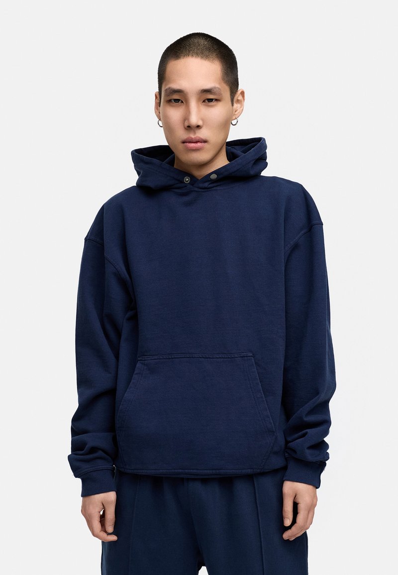 Marineblaue Kapuzenjacke aus Baumwolle, mit einer Fronttasche, Kordelzug-Kapuze und gerippten Bündchen. Lockerer Schnitt mit überschnittenen Schultern.