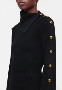 Gros plan d’une personne portant un pull en tricot noir avec un col asymétrique haut et des boutons dorés le long de l’épaule et de la manche.