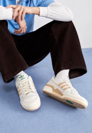 Osoba siedząca z założonymi skrzyżowanymi ramionami, ubrana w kremowe i beżowe buty Adidas, białe skarpetki, ciemnobrązowe spodnie oraz niebieską koszulę, na jasnoniebieskiej podłodze.