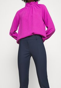 Fuchsiová halenka s dlouhým rukávem a nařaseným výstřihem v uvolněném střihu, spárovaná s tmavě modrými elegantními kalhotami s hladkou texturou a štíhlými nohavicemi.