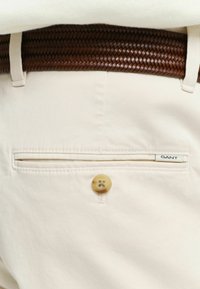 Beige katoenen broek met een achterzak die een bruine knoop en een GANT-label heeft. De tailleband is voorzien van een gevlochten bruine leren riem.
