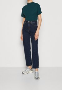 Tmavě modré džíny s vysokým pasem v kombinaci s krátkým rukávem tyrkysovým topem a stříbrnými sportovními botami, disponující volným střihem a minimalistickým designem.