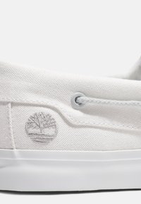 White canvas sneaker featuring a textured finish, round eyelets, and a grey embroidered tree logo on the side. Midsole in solid white.