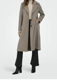 Abrigo de lana marrón claro con solapas anchas, cierre de un solo botón y bolsillos laterales. Combinado con pantalones negros acampanados y botas de tacón.