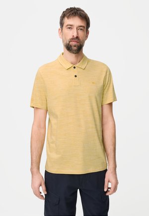 Mann mit kurzen braunen Haaren und Bart, der ein hellgelbes Poloshirt und dunkle Hose trägt, vor einem schlichten weißen Hintergrund stehend.