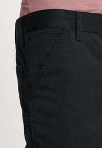 Czarne spodnie o gładkiej teksturze, prostym kroju i tradycyjnym fasonie z pięcioma kieszeniami, szlufkami na pasek i zamkiem błyskawicznym.