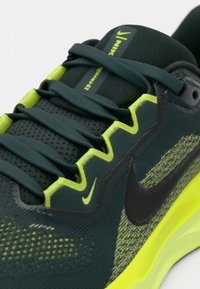 Κοντινή λήψη από αθλητικά παπούτσια Nike σε σκούρο πράσινο και νέον κίτρινο χρώμα με μαύρο σήμα και χονδρές κορδόνες, κατασκευασμένα από αναπνεύσιμο δίχτυ.