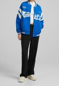 Niebieska sportowa kurtka z białymi napisami i paskami, z zapięciem na guziki i luźnym krojem, połączona z czarnymi spodniami i beżowymi sneakersami.