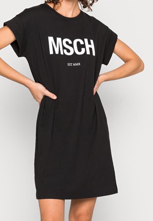 Femme portant une robe noire à manches courtes avec les inscriptions "MSCH" et "EST MMX" en blanc, mains sur les hanches, debout devant un fond uni.