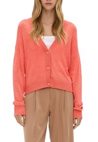 Koralowy dzianinowy kardigan z dekoltem w serek, ściągaczami na rękawach i guzikami z przodu. W połączeniu z jasnobrązowymi plisowanymi spodniami. Miękka faktura i luźny krój.